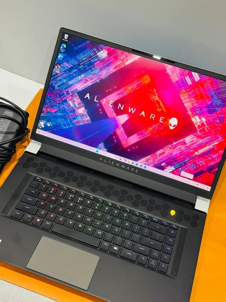 A close-up, angled view testing unit of an Alienware gaming laptop showing its bright screen, multi-colored RGB keyboard, trackpad, and the distinct honeycomb speaker grill above the keys, with a power adapter visible to the left.